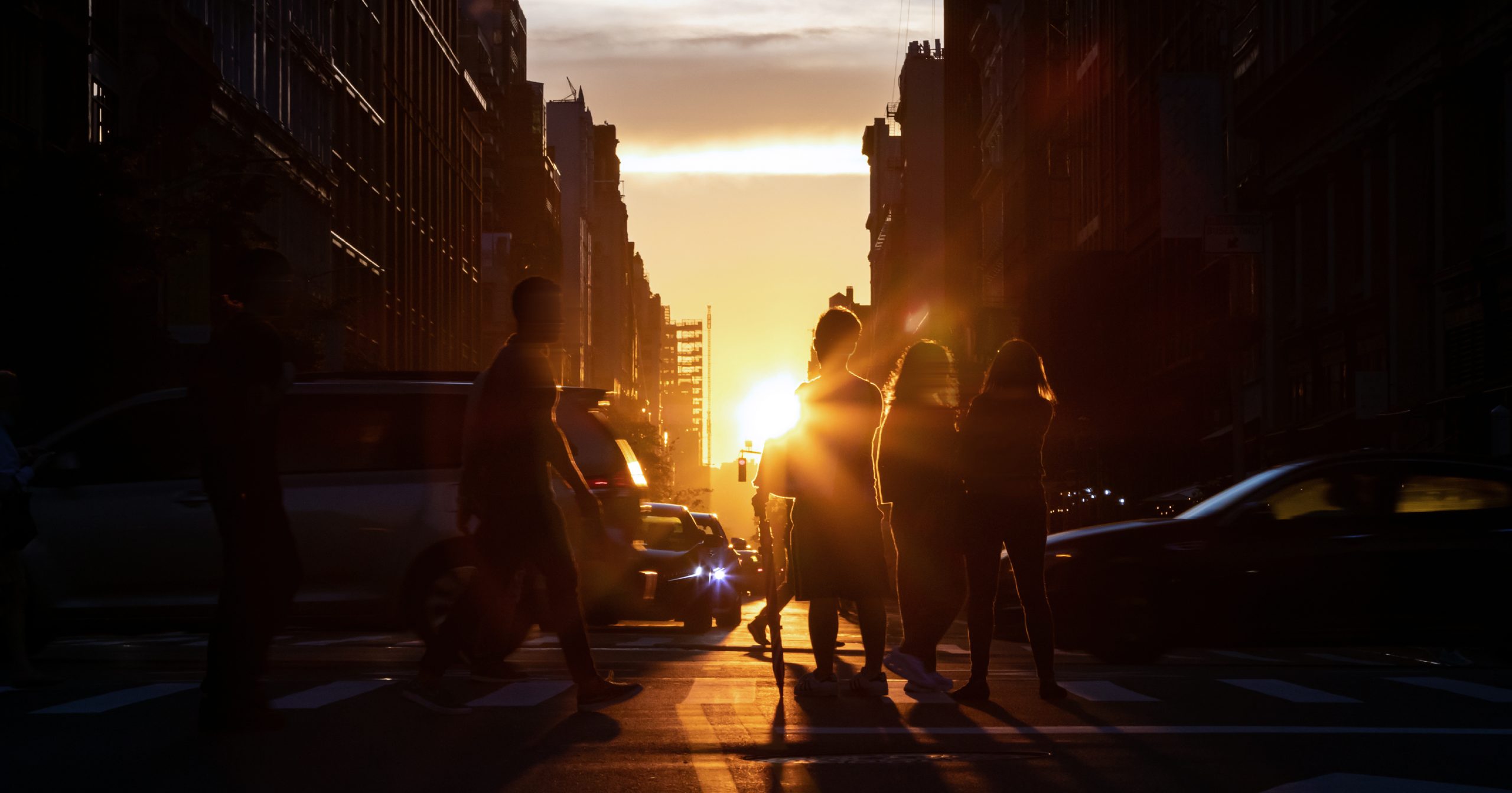Image shows a crowded intersection with the silhouette of cars and pedestrians crossing, while the sun sets in the background.
